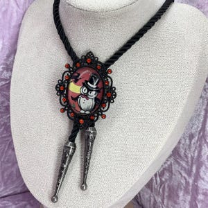 May include: Black bolo tie with an oval pendant featuring an owl in a top hat against a red background. The pendant is framed with a black filigree and red rhinestones. The tie has a black cord and silver-coloured tips.