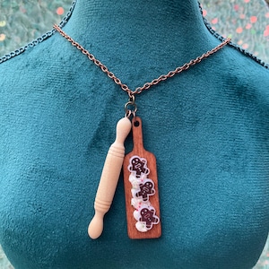 May include: A copper chain necklace with a pendant featuring a miniature rolling pin and a wooden cutting board. The cutting board has three gingerbread men with white icing and sprinkles. The rolling pin and cutting board are made of wood.