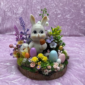May include: A white ceramic bunny figurine holding a brown egg, surrounded by pastel colored Easter eggs and spring flowers in a wooden planter.