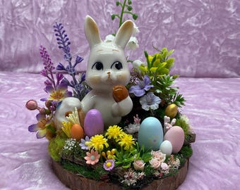 May include: A white ceramic bunny figurine holding a brown egg, surrounded by pastel colored Easter eggs and spring flowers in a wooden planter.