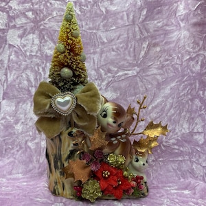 Könnte beinhalten: Eine Keramik-Weihnachtsbaumfigur mit einer Samtschleife und einem herzförmigen Ornament. Zwei Keramik-Hirschfiguren sind am Fuß des Baumes eingebettet, umgeben von goldenem und grünem Laub.