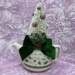 May include: A white teapot with a green velvet bow and a small white flocked Christmas tree on top. The teapot has a holly and berry design.