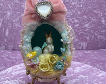 May include: A vintage Easter egg decoration with a pink velvet bow and a rhinestone heart. Inside the egg, a porcelain bunny sits on a bed of yellow and blue flowers.