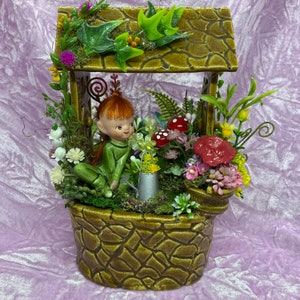 May include: A ceramic fairy figurine with green clothing and red hair sits in a brown and green planter with a stone-like texture. The planter is filled with artificial flowers and greenery, including red mushrooms and a small watering can.