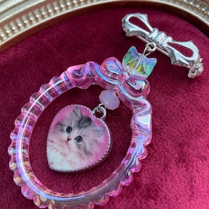 Puede incluir: Broche decorativo con un marco rosa e iridiscente y un colgante en forma de corazón. El colgante muestra la cara de un gatito esponjoso. Se adjunta un alfiler plateado en forma de lazo. Una pequeña perla rosa cuelga del marco.