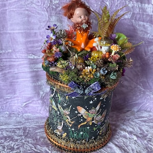 Puede incluir: Una pieza decorativa cilíndrica y caprichosa con una figura de muñeca sobre un lecho de plantas, flores y setas artificiales. La base es un cilindro negro adornado con ilustraciones de hadas y el texto "FULL MOON". Adornado con abalorios dorados y negros.