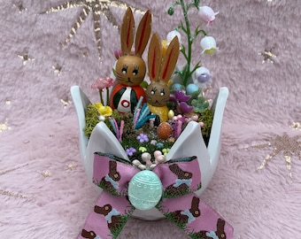 May include: A white ceramic planter shaped like a flower with a pink ribbon tied around it. The planter is filled with moss, small flowers, and two wooden bunny figurines. The bunny figurines are painted with brown, yellow, and red details. The ribbon has a repeating pattern of brown bunnies.