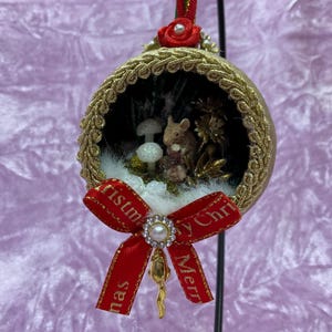May include: A gold Christmas bauble with a miniature scene inside. The scene features a small mouse, mushrooms, and greenery. The bauble is decorated with a red ribbon with the words "Merry Christmas" written on it.