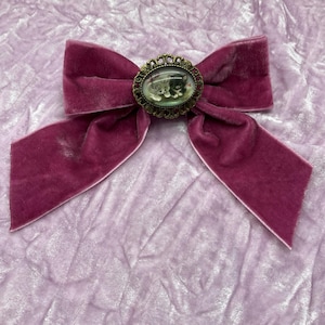 May include: A pink velvet bow with a gold-toned cameo featuring two kittens. The cameo is oval-shaped and has a detailed floral design around the edge.