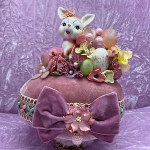 May include: A pink velvet Easter egg with a white bunny figurine, pink and green floral accents, and a pink bow.