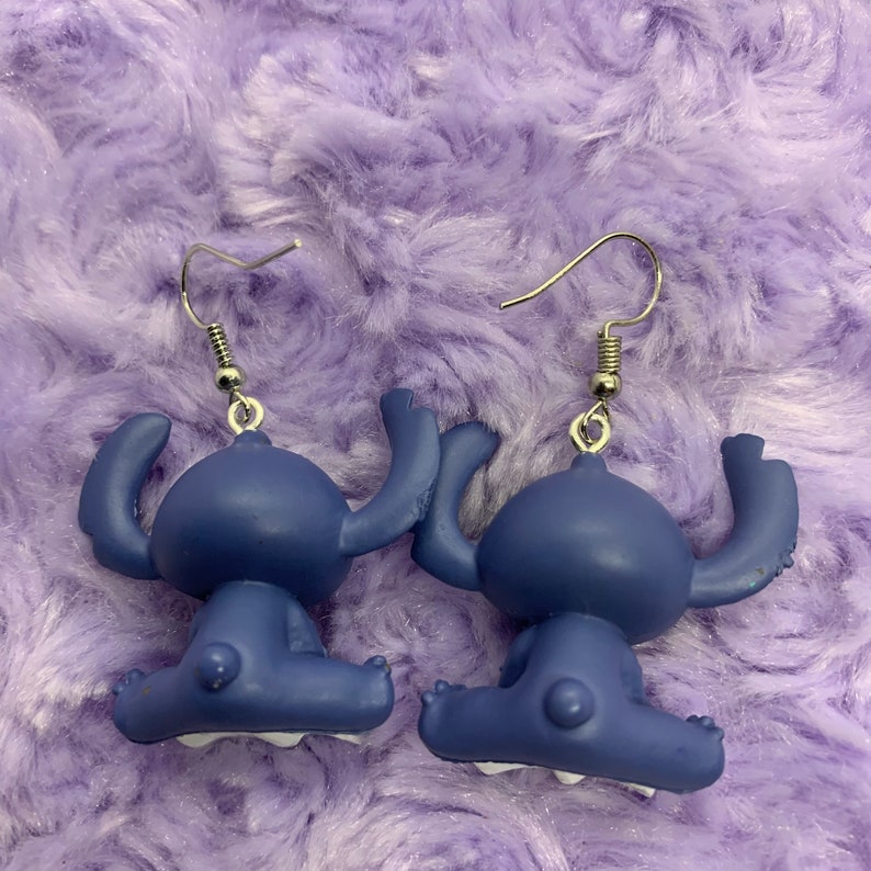 Puede incluir: Un par de pendientes colgantes azules con el personaje de Stitch y ganchos plateados. Los pendientes est&aacute;n hechos de pl&aacute;stico y presentan un dise&ntilde;o 3D de Stitch sentado con los brazos levantados.