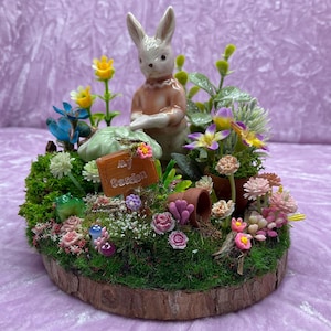 May include: A miniature garden scene with a ceramic bunny figurine, a green mushroom, a sign that says "My Garden", and various flowers and plants. The scene is set on a wooden slice.