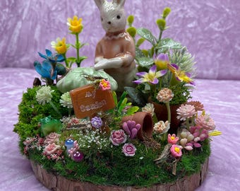 May include: A miniature garden scene with a ceramic bunny figurine, a green mushroom, a sign that says "My Garden", and various flowers and plants. The scene is set on a wooden slice.
