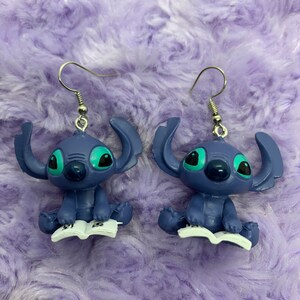 Puede incluir: Un par de pendientes azules de Stitch con ojos verdes. Stitch sostiene un libro en sus manos. Los pendientes son plateados y tienen un cierre de gancho.