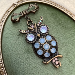 May include: A bronze-toned owl brooch with iridescent blue and clear crystal accents. The owl has large, round eyes and is adorned with multiple crystals. The brooch is attached to a bow-shaped clasp.