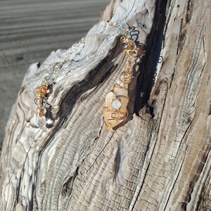 May include: A gold wire wrapped pendant with a clear and peach colored crystal. The pendant is hanging from a gold chain on a piece of driftwood.