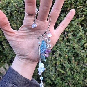 May include: A silver ear cuff with a dangling charm featuring iridescent blue and purple stones, clear beads, and a silver chain.