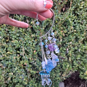 May include: Silver ear cuff with a dangling jellyfish charm and iridescent beads in shades of blue, purple, and white.