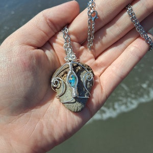 May include: A silver chain necklace with a pendant made from a grey rock with a blue and green iridescent stone set in the center. The rock is wrapped in silver wire.