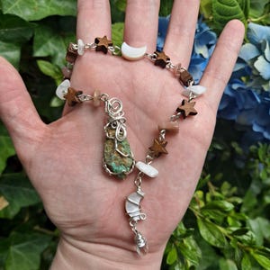 May include: A handmade necklace featuring a pendant with a turquoise stone wrapped in silver wire. The necklace includes a chain with wooden stars, moon-shaped shell, and various gemstones. The background is a mix of green and blue foliage.