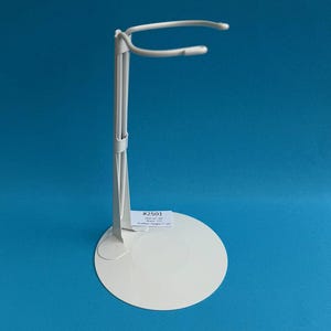 May include: A white metal doll stand with a circular base and a curved top. The stand has a vertical support and a small label with text. The stand's overall height is 17.78 cm.
