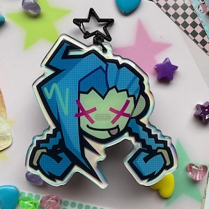 以下が含まれることがあります： ピンクのXが目に描かれた青と緑のキャラクター、青と緑のピクセルデザイン、青と緑のピクセルヘアスタイル。キャラクターは、銀色の星チャームが付いた透明なアクリルキーホルダーに付いています。