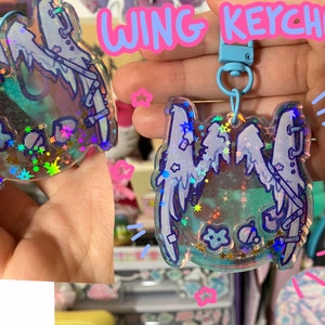 May include: A clear, wing-shaped keyring with a blue clasp. The keyring features a blue outline with a wing design, filled with iridescent glitter and small star-shaped confetti. The text "WING KEYCHAIN!" is above the keyring.