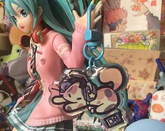 May include: A clear keychain with a cartoon cat wearing headphones and a backpack. The cat is light brown with dark brown spots. The keychain is attached to a blue metal ring.