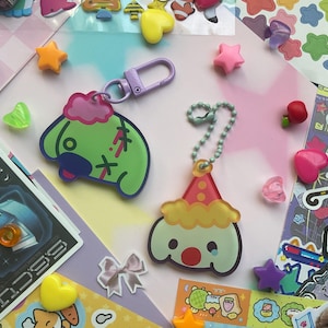 May include: Two colourful keychains featuring cartoon dog characters. One keychain is green with a pink hat and stitches, the other is yellow with a red and white party hat. Both keychains have black eyes and a red nose.