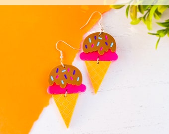 May include: A pair of ice cream cone earrings with pink, brown, and white details. The earrings are made of acrylic and are perfect for summer.