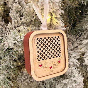 Op de afbeelding: Een houten kerstornament in de vorm van een radio met een rode zijkant en een lichthouten voorkant met een roosterpatroon. De voorzijde is gegraveerd met de namen Paul Age 3 en Jessi Age 6, het jaartal 2026 en kleine rode hartjes.