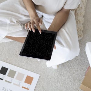May include: A person is holding a black tablet with a white dot pattern. A color palette with black, gray, and beige swatches is on the floor. A white box with pastries and a brown box are also present.