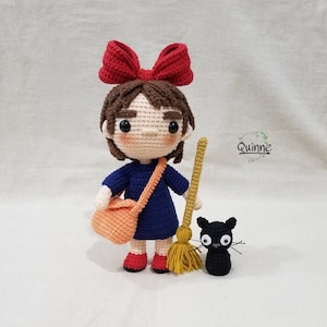 May include: A crocheted doll with brown hair, a red bow, and a blue dress holding a broom and a black cat. The doll is wearing a red and orange bag.