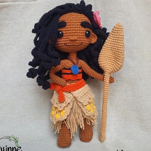 May include: Crocheted Moana doll with brown skin, black curly hair, and a blue necklace. She is wearing a red and orange top, a brown skirt with yellow flowers, and holding a brown wooden spear. The doll is standing on a beige fabric background.