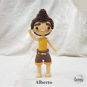 Puede incluir: Una muñeca de crochet con cabello castaño, un top amarillo sin mangas, pantalones cortos marrones y una expresión amigable. La muñeca se llama "Alberto".