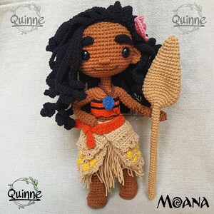 May include: Crocheted Moana doll with black hair, brown skin, and a blue and orange top. She is holding a brown wooden paddle. The doll is standing on a beige surface. The text "Moana" is written in a stylized font below the doll.