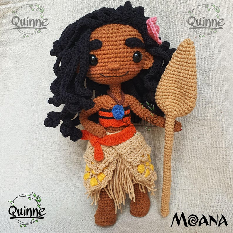 May include: Crocheted Moana doll with black hair, brown skin, and a blue and orange top. She is holding a brown wooden paddle. The doll is standing on a beige surface. The text "Moana" is written in a stylized font below the doll.