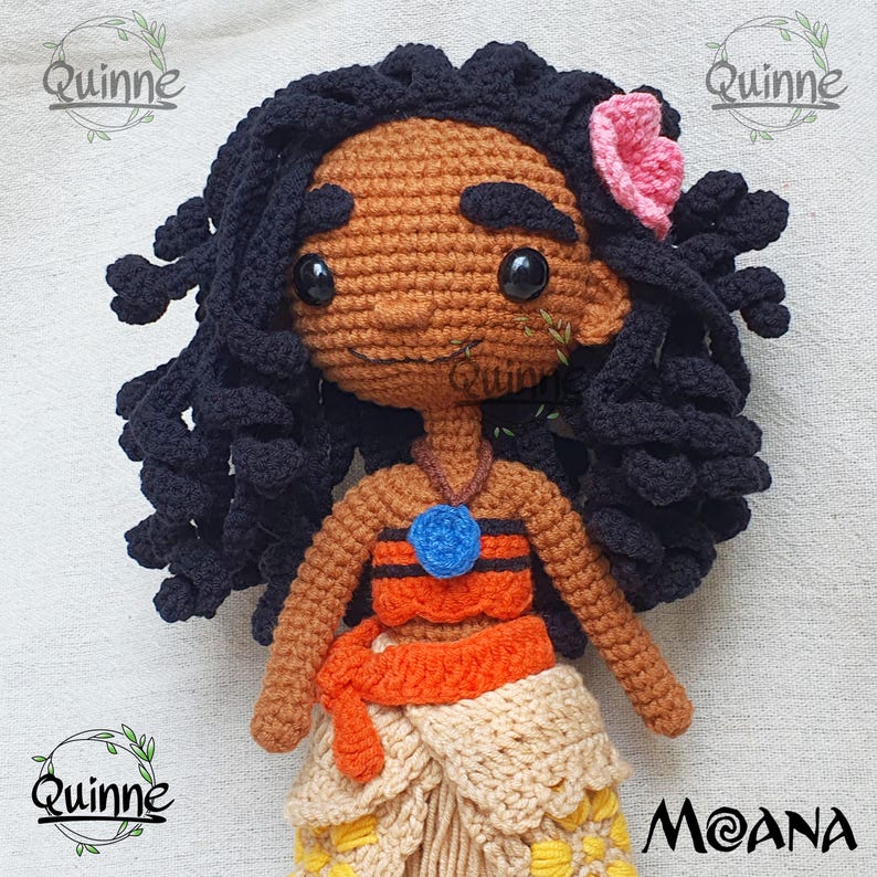 May include: Crocheted Moana doll with black hair, brown skin, and a flower behind her ear. She is wearing a brown and orange top, a blue necklace, and a brown and orange skirt.