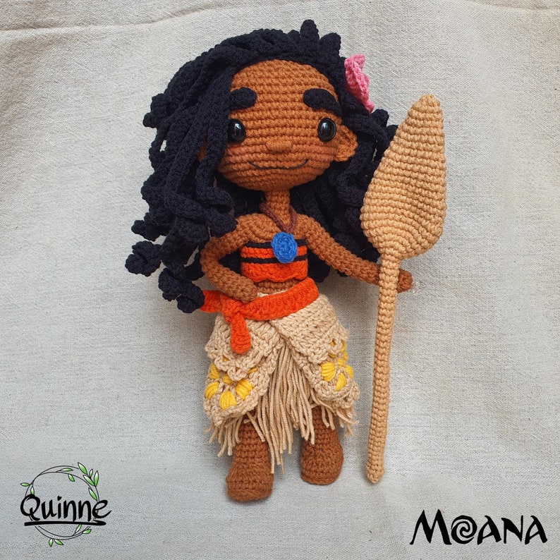 May include: Crocheted Moana doll with brown skin, black curly hair, and a blue necklace. She is wearing a red and orange top, a brown skirt with yellow flowers, and holding a brown wooden spear. The doll is standing on a beige fabric background.