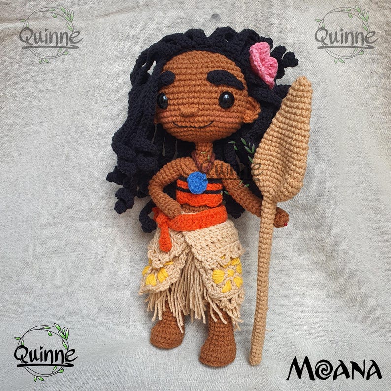 May include: Crocheted Moana doll with brown skin, black hair, and a pink flower. She is wearing a brown and orange outfit and holding a brown stick.