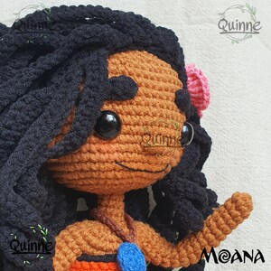 May include: Crocheted Moana doll with brown skin, black hair, and a blue necklace. The doll is wearing a brown top and a pink flower in her hair. The doll is smiling and has a friendly expression.
