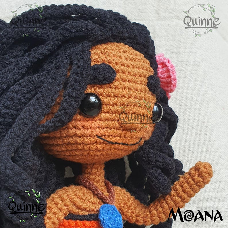 May include: Crocheted Moana doll with brown skin, black hair, and a blue necklace. The doll is wearing a brown top and a pink flower in her hair. The doll is smiling and has a friendly expression.