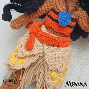 May include: Crocheted Moana doll with brown skin, black hair, and a tan and orange outfit. The outfit features a tan skirt with yellow flower accents and an orange sash. The doll is wearing a blue necklace with a blue flower pendant.