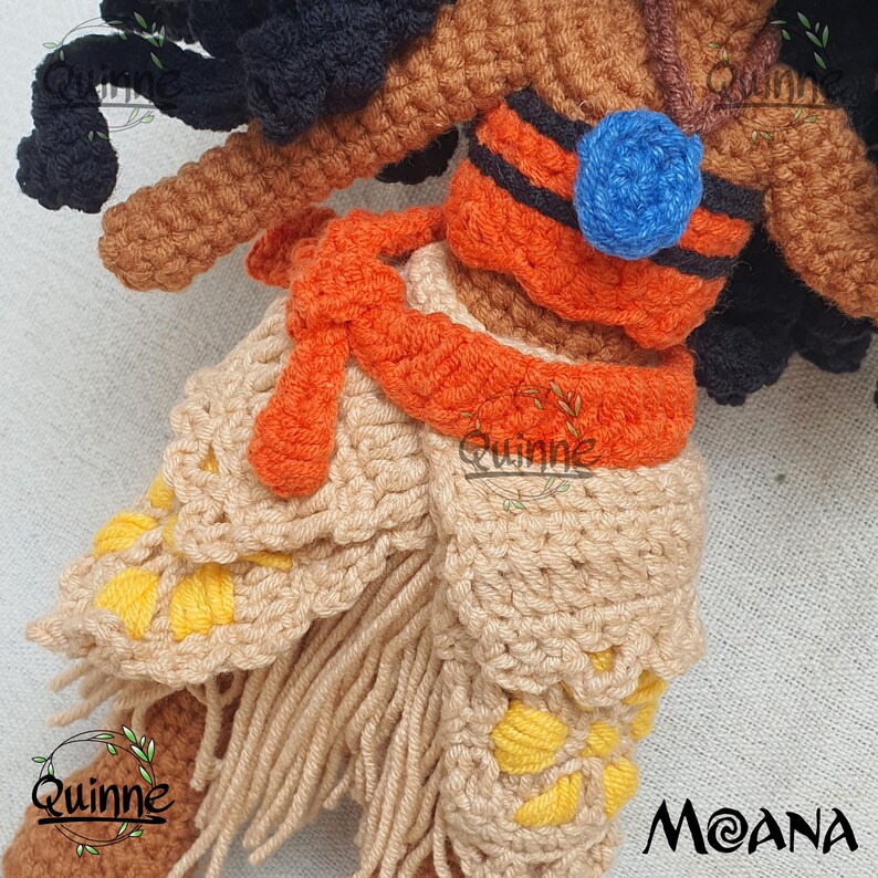 May include: Crocheted Moana doll with brown skin, black hair, and a tan and orange outfit. The outfit features a tan skirt with yellow flower accents and an orange sash. The doll is wearing a blue necklace with a blue flower pendant.