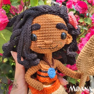 May include: Crocheted Moana doll with brown skin, black hair, and a blue necklace. She is wearing an orange and brown skirt and holding a brown spear. The doll is surrounded by pink flowers.