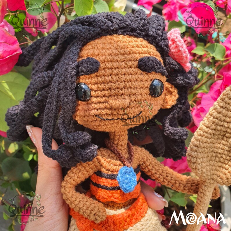 May include: Crocheted Moana doll with brown skin, black hair, and a blue necklace. She is wearing an orange and brown skirt and holding a brown spear. The doll is surrounded by pink flowers.