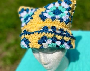May include: A crocheted hat with a yellow, blue, white, and green geometric pattern. The hat has two pointed ear flaps.
