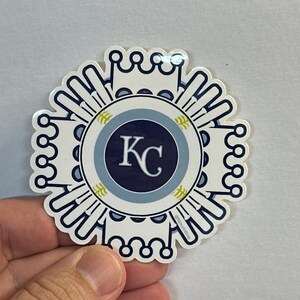 Pode incluir: Um adesivo branco com um design azul e dourado. O centro do adesivo apresenta um c&iacute;rculo azul com um contorno branco e as letras "KC" em branco. O c&iacute;rculo &eacute; rodeado por um anel branco com um contorno azul e um design de folhas douradas.