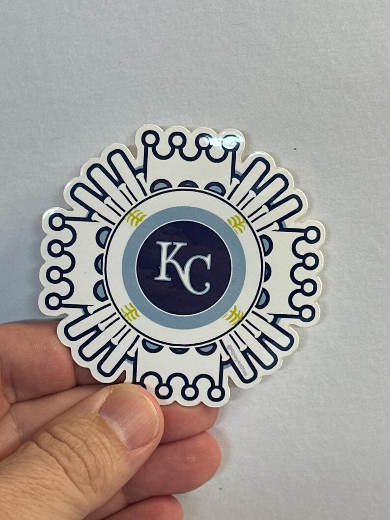 Pode incluir: Um adesivo branco com um design azul e dourado. O centro do adesivo apresenta um c&iacute;rculo azul com um contorno branco e as letras "KC" em branco. O c&iacute;rculo &eacute; rodeado por um anel branco com um contorno azul e um design de folhas douradas.