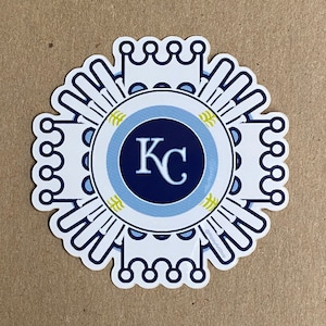 Pode incluir: Etiqueta branca com um design circular azul e azul claro. O centro do c&iacute;rculo tem um c&iacute;rculo azul marinho com as letras "KC" em branco. A etiqueta tem um design de coroa ao redor da borda externa.